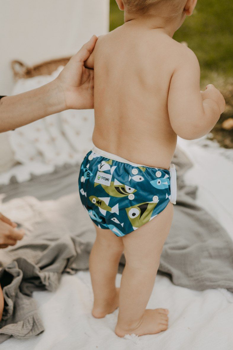 Baby trägt eine Schwimmwindel mit blauem Fischmuster, T-TOMI Logo sichtbar.