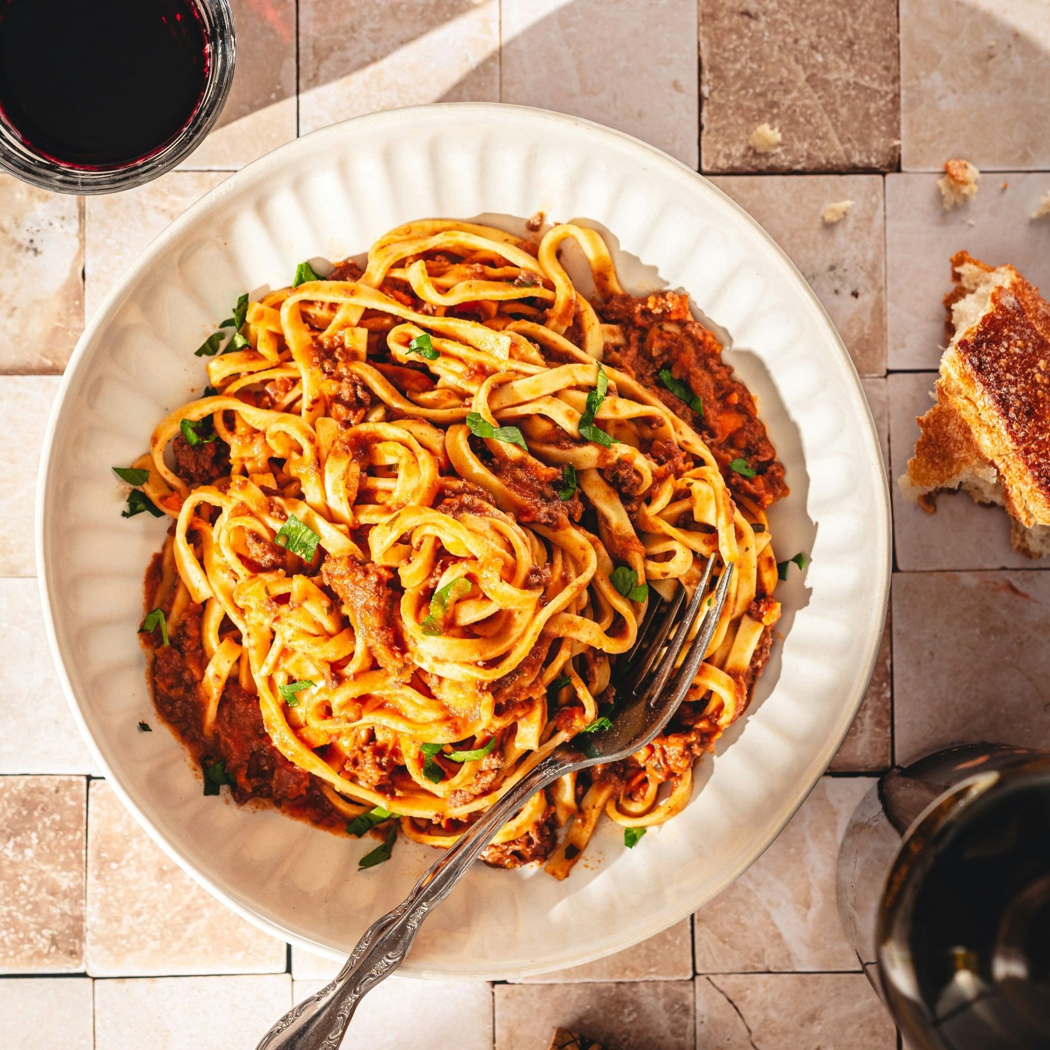 Teller mit Pasta Bolognese, Gabel. Brot und Weinglas im Hintergrund. Auf quadratischen Fliesen.