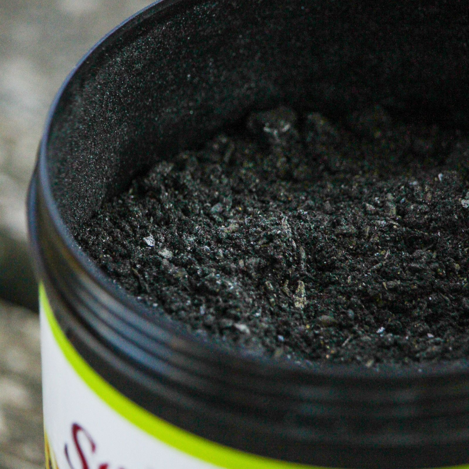 Geöffnete Dose mit schwarzem Pulver. Schwarzer Behälter mit grünem Rand. Produktname nicht vollständig sichtbar.