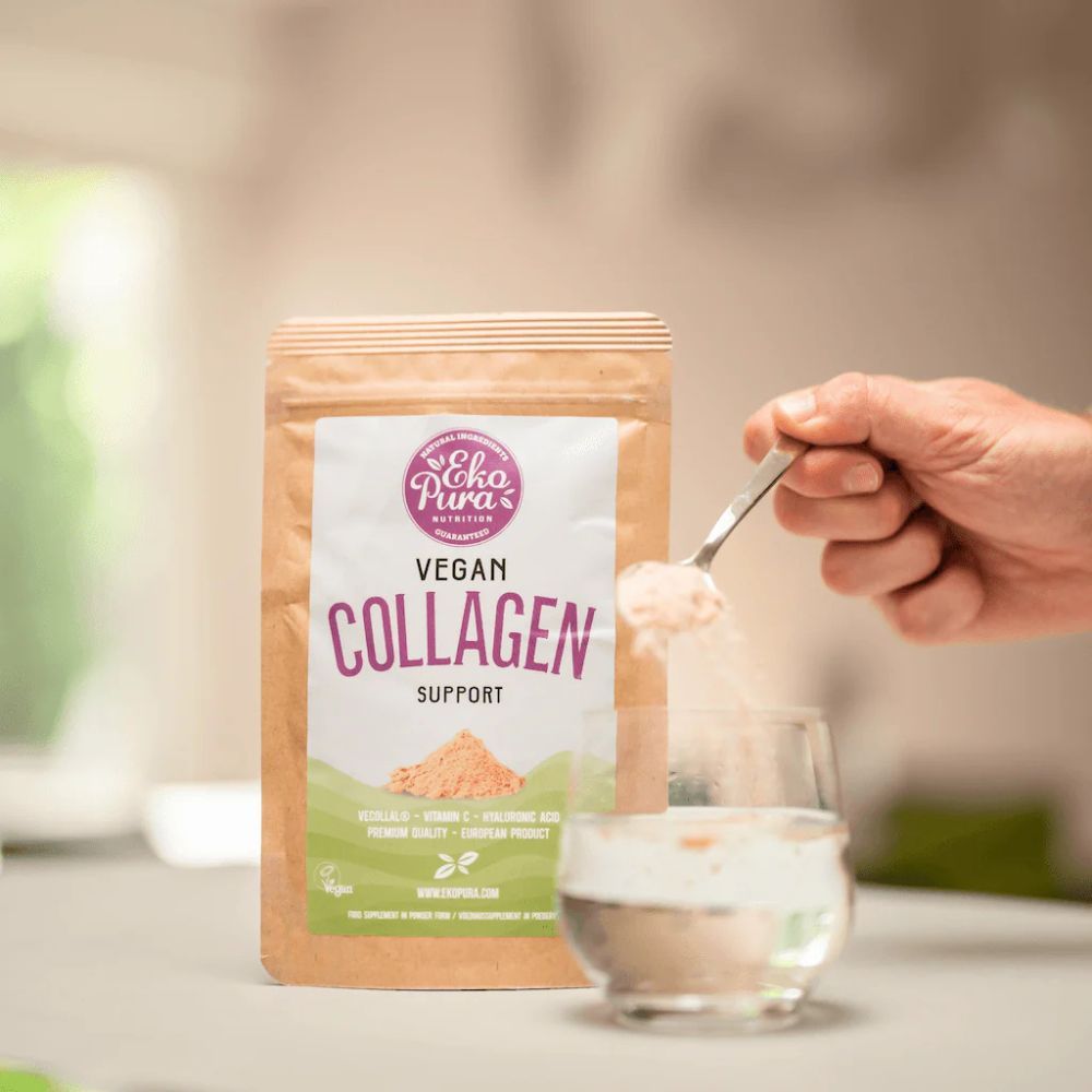 Beutel mit Aufschrift Vegan Collagen Support. Löffel mit Pulver über Glas. Hand hält Löffel. Hintergrund: Küche.