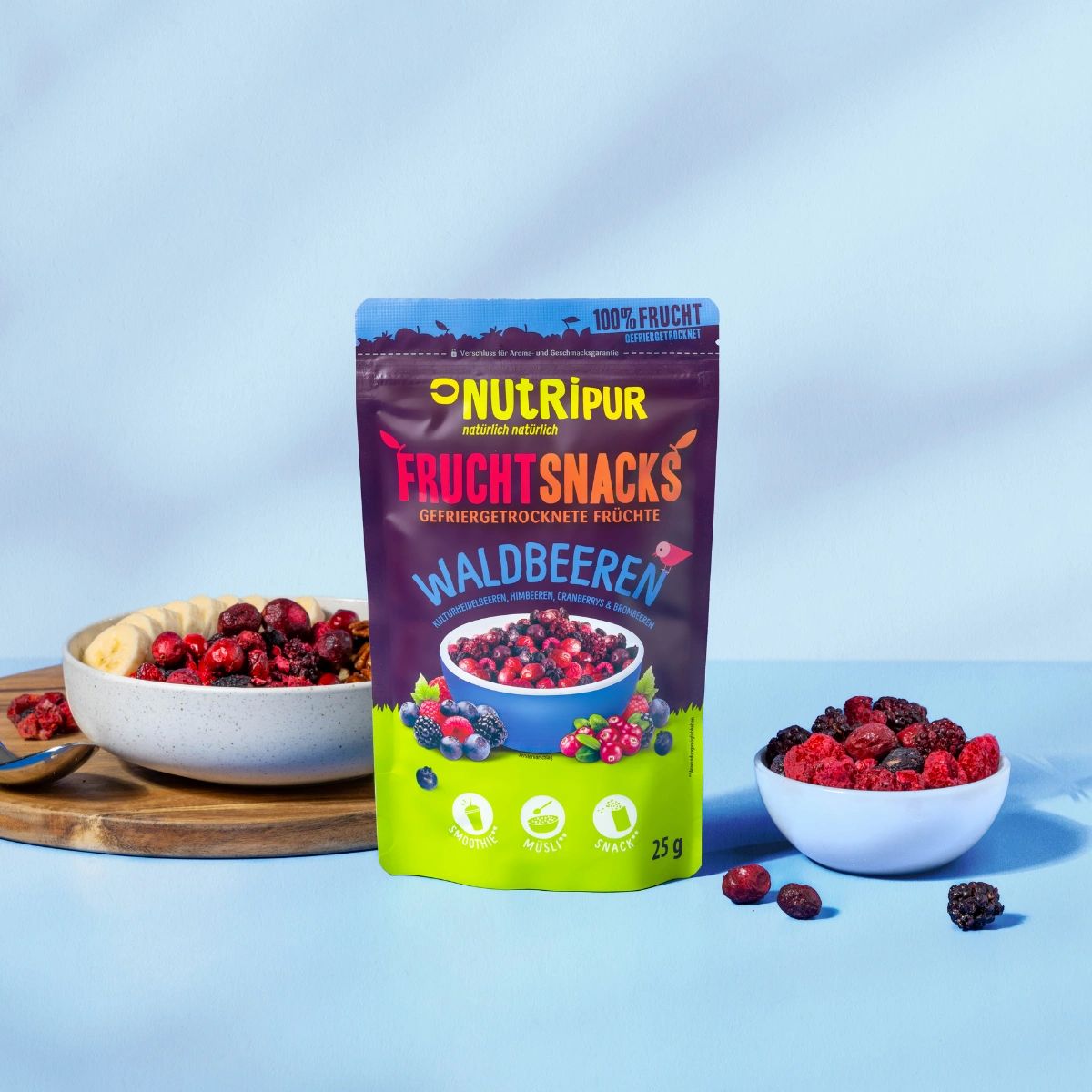 Beutel NutriPur Frucht-Snacks Waldbeeren, daneben Schüssel mit Beeren und Bananenscheiben. Auf blauem Hintergrund.