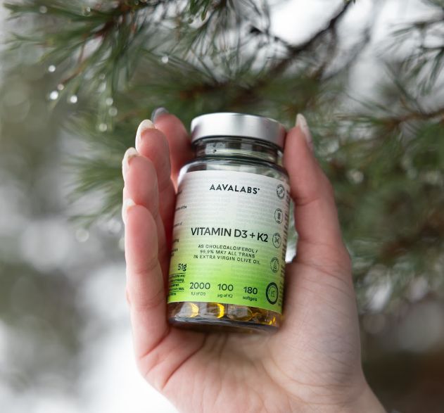 Eine Hand hält eine Flasche Aavalabs Vitamin D3+K2 Softgels vor einem verschwommenen Hintergrund aus Tannenzweigen.