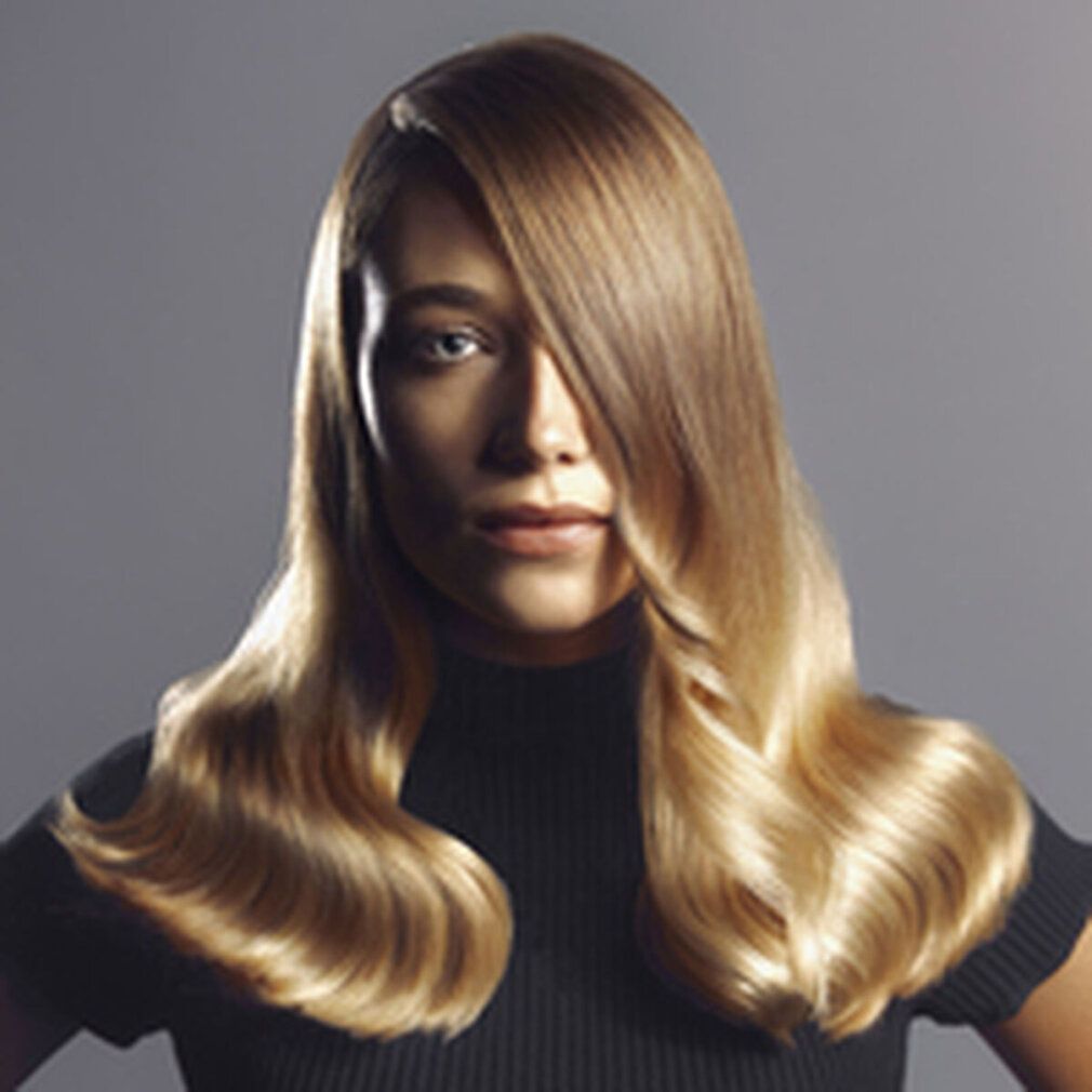 Porträt einer Frau mit blondem, gewelltem Haar. Sie trägt ein schwarzes Oberteil. Haare fallen über eine Schulter.