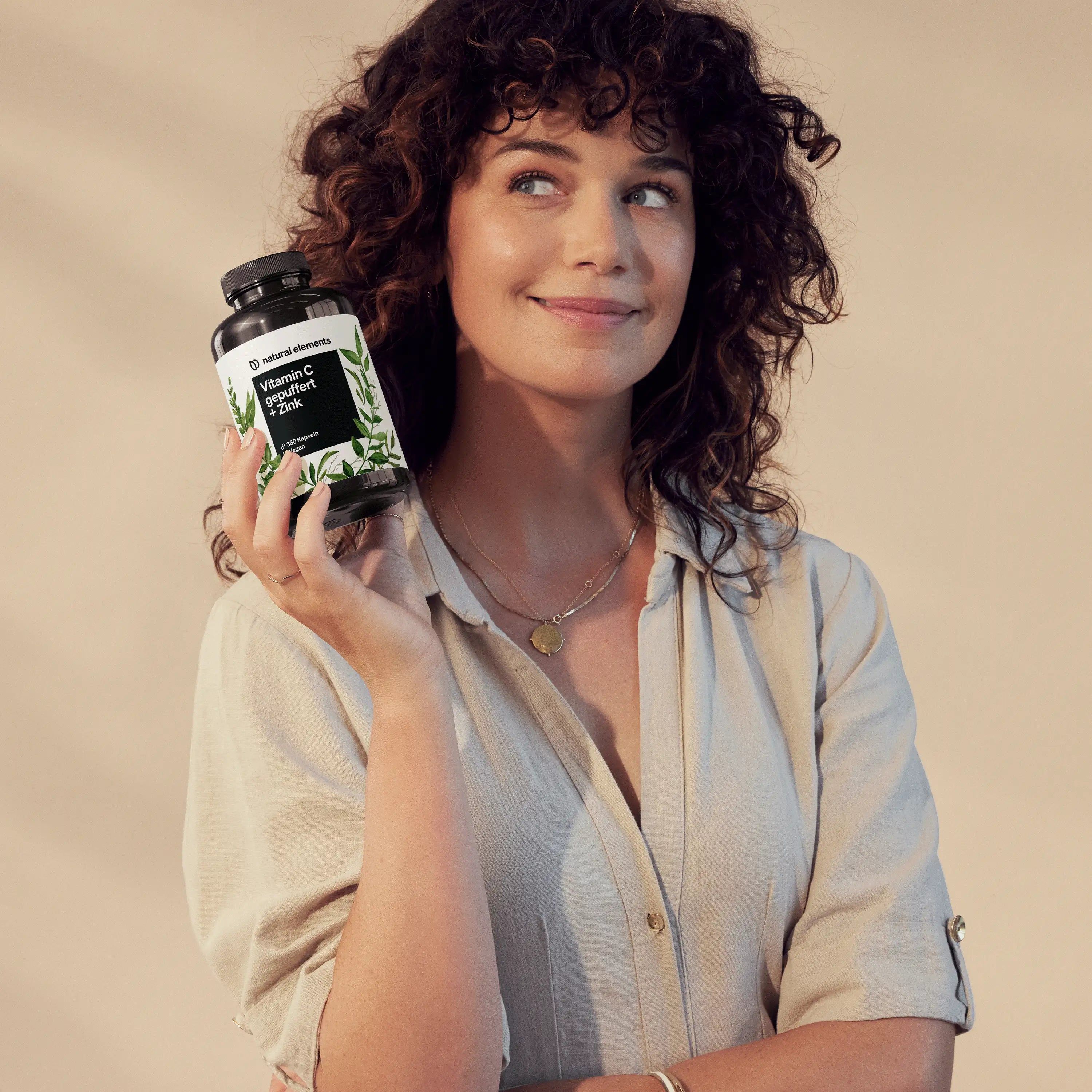 Frau hält eine schwarze Flasche mit weißem Etikett. Aufschrift: Vitamin C gepuffert + Zink. Marke: natural elements. Frau lächelt.