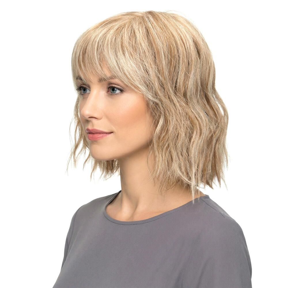Blond gewellte Perücke mit Pony. Seitenansicht einer Frau mit grauer Bluse. Der Hintergrund ist weiß.