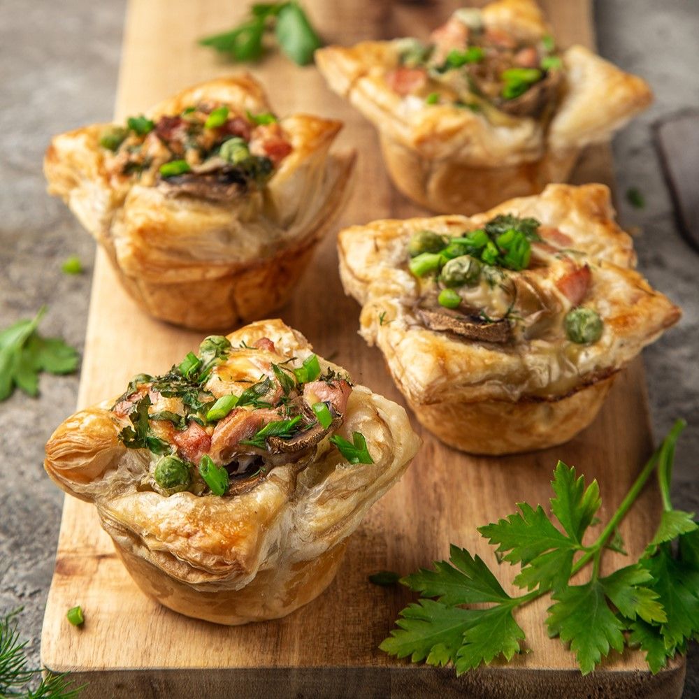 Vier herzförmige Blätterteig-Tartelettes mit Pilzen, Erbsen und Kräutern auf Holzbrett.