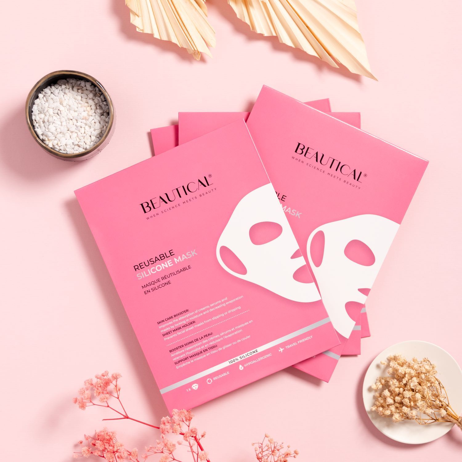 Plusieurs emballages roses avec masque en silicone blanc. Texte: BEAUTICAL, REUSABLE SILICONE MASK. Décoration: fleurs séchées, bol.