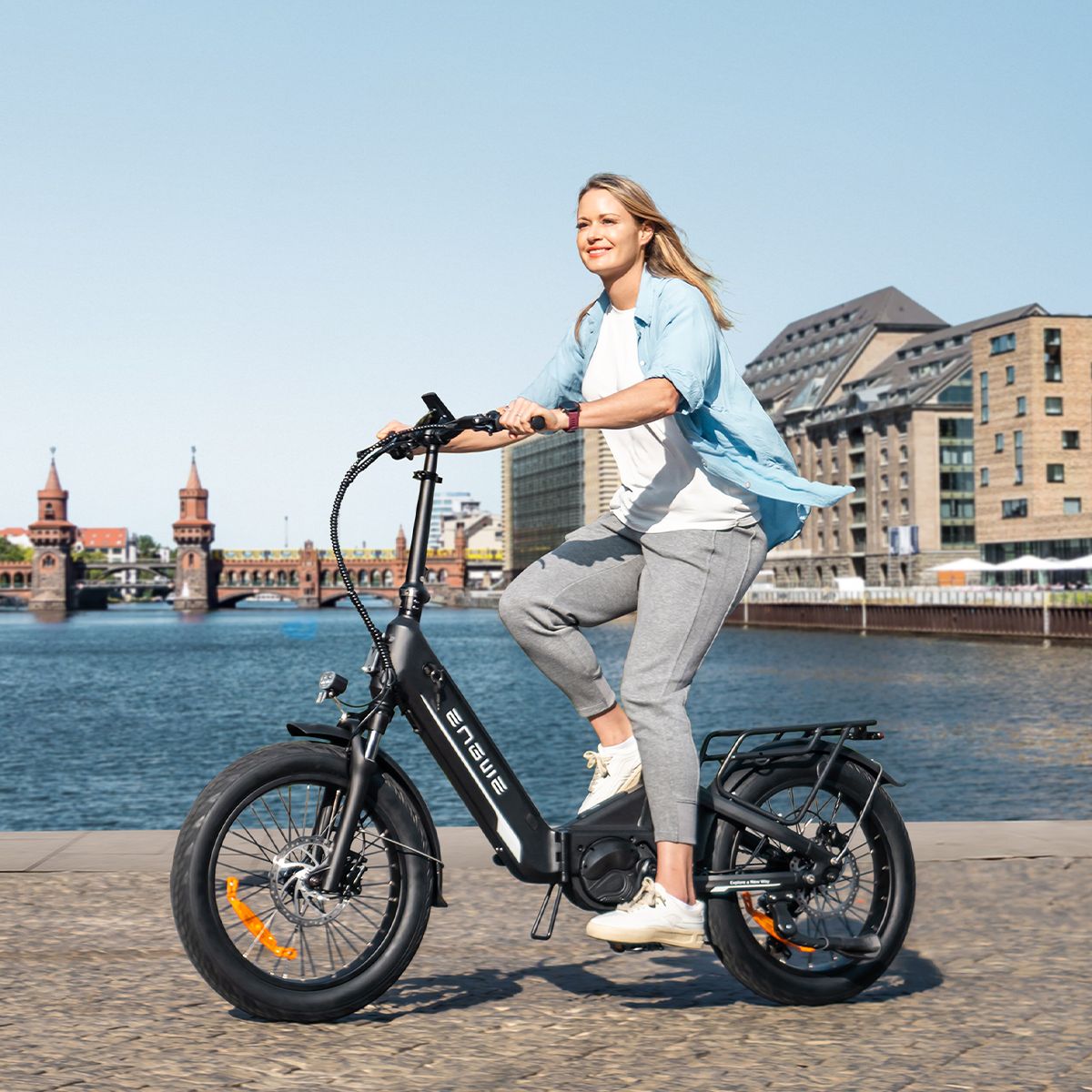 Frau fährt auf einem schwarzen ENGWE L20 3.0 Pro E-Bike. Hintergrund: Wasser und Gebäude.