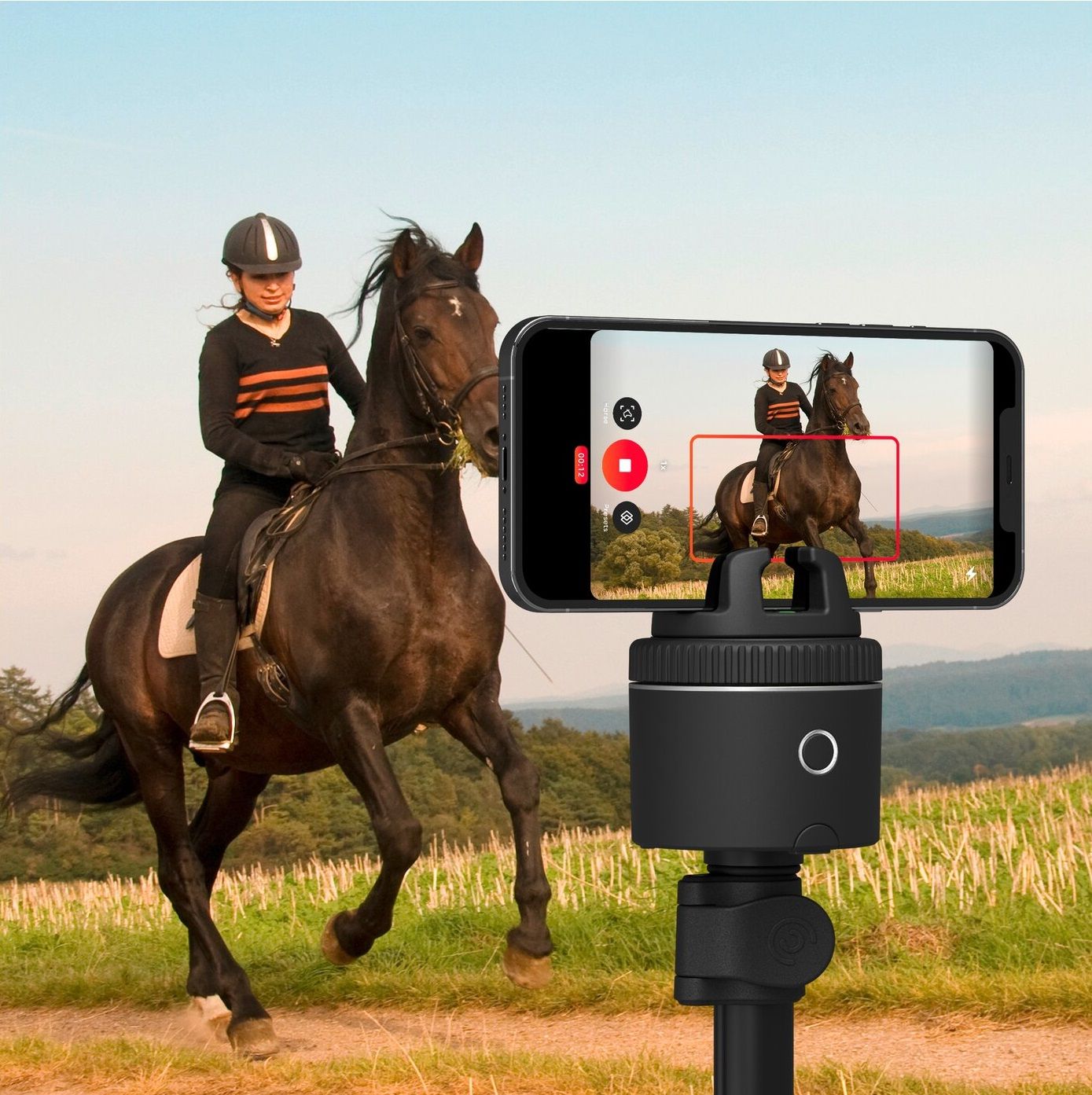 Pivo-Stativ mit Smartphone, das eine Reiterin auf einem Pferd filmt. Bildschirm zeigt Aufnahme. Grünfläche im Hintergrund.
