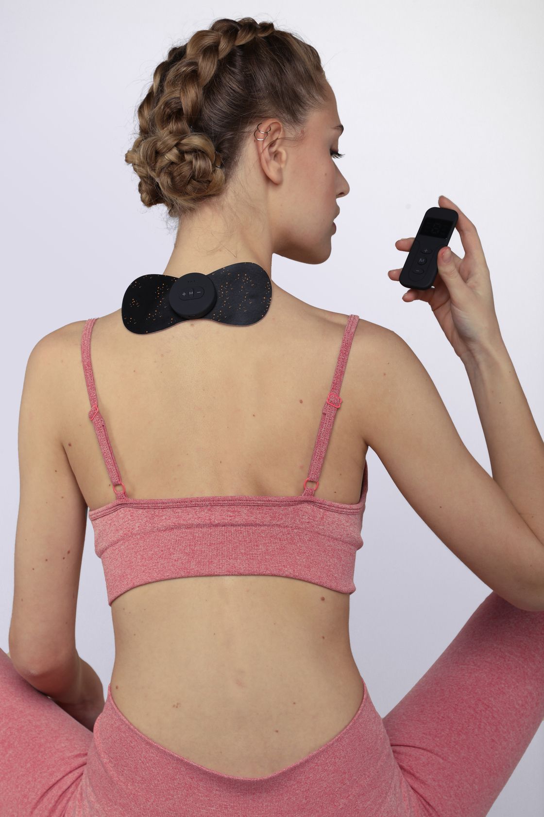 Person mit Massagepad am Nacken, hält Fernbedienung. Schwarzes Pad mit blauem Rand und digitalem Display.