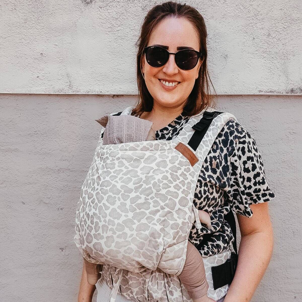 Frau mit Sonnenbrille trägt Babytrage mit Leopardenmuster. Baby im Tragetuch.