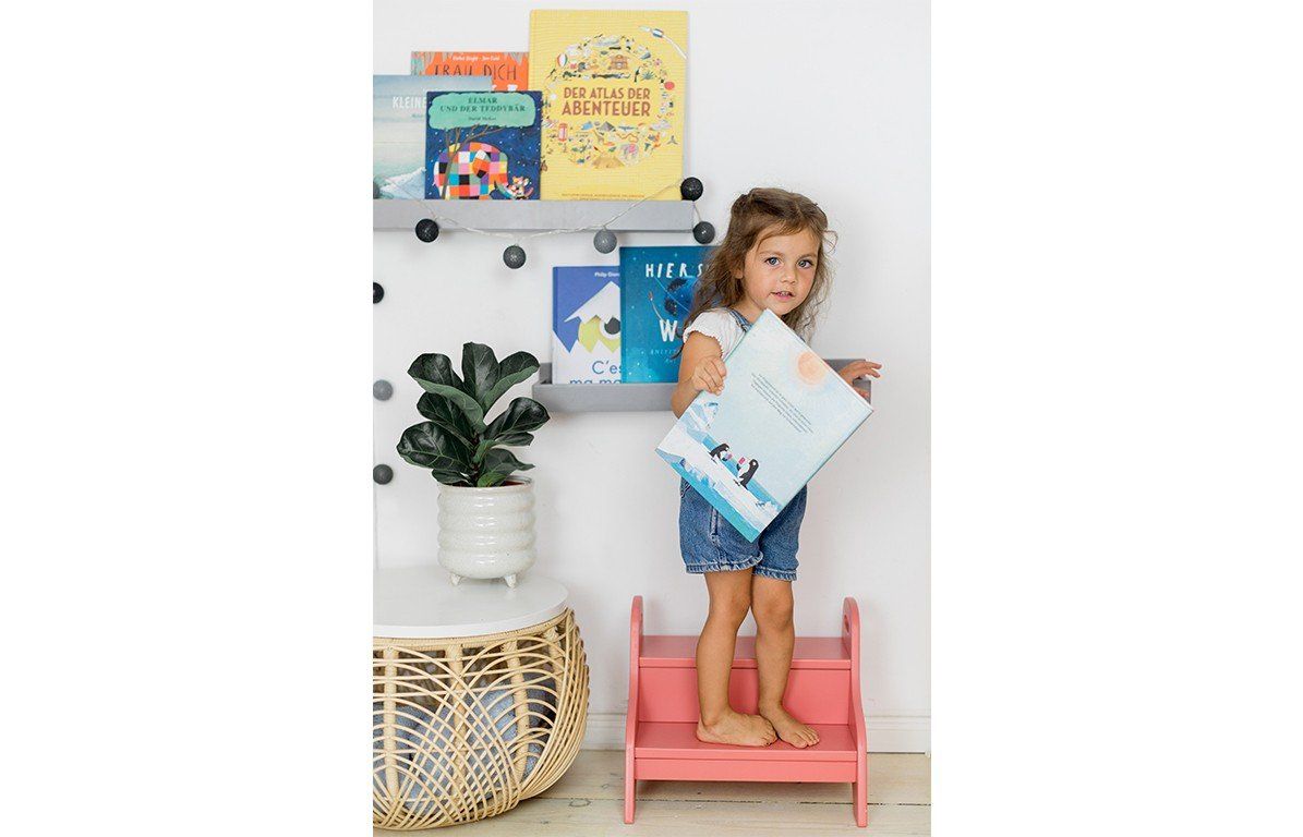 Kind steht auf einem Holztritt, altrosa, zweistufig. Bücherregal im Hintergrund.