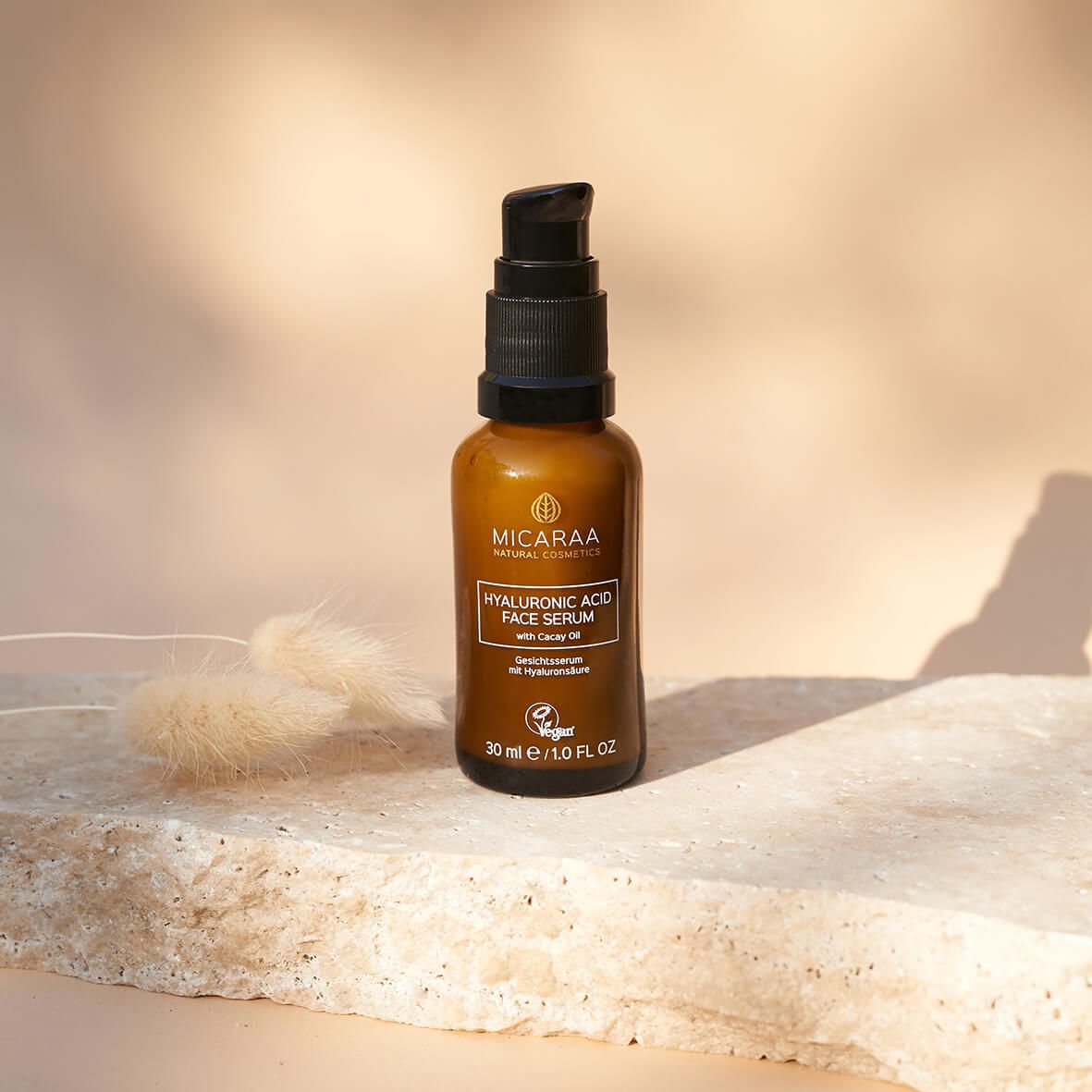 Braune Glasflasche mit schwarzem Pumpverschluss. Aufschrift: MICARAA Hyaluronic Acid Face Serum. Auf Stein platziert.