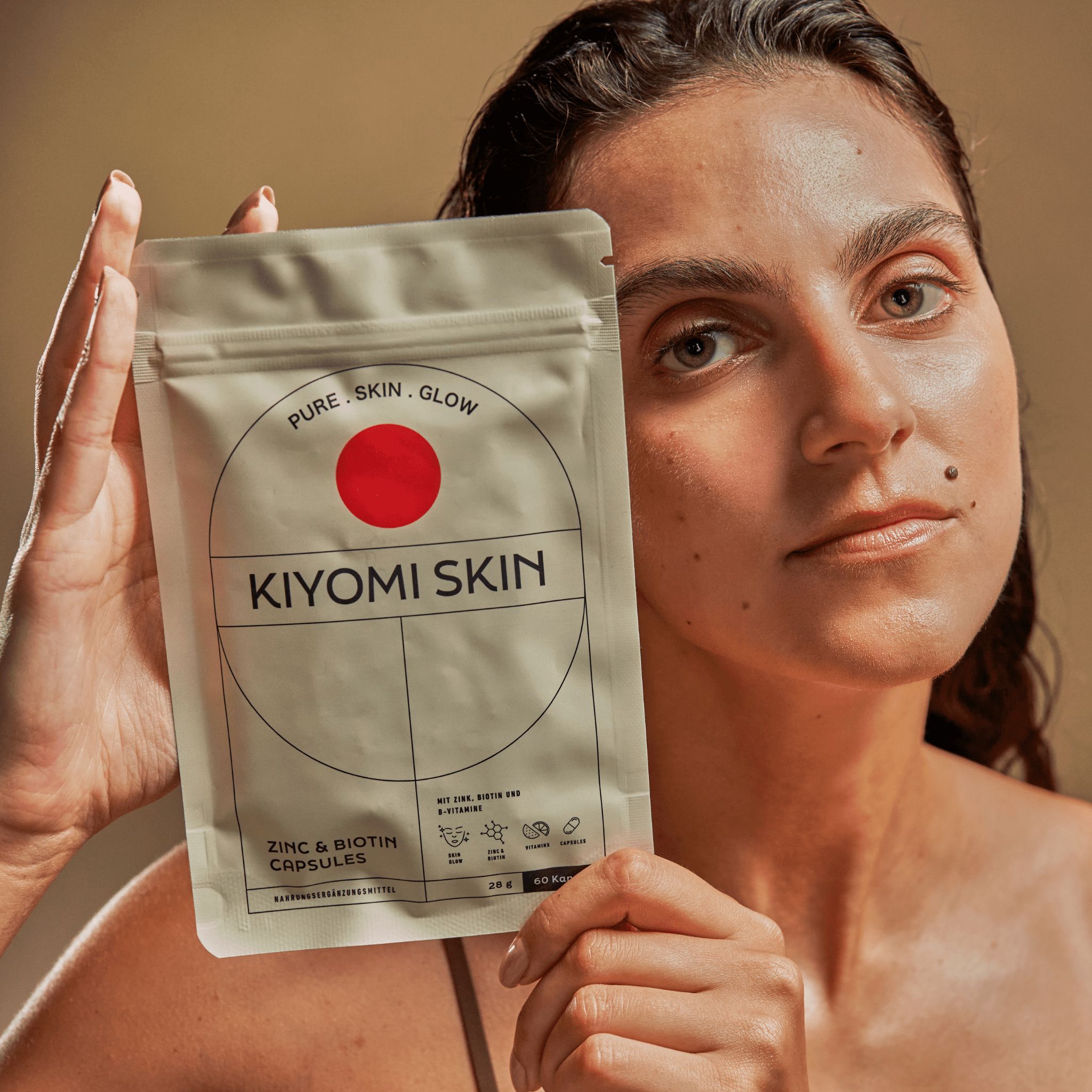 Frau hält Kiyomi Skin Glow Capsules-Beutel. Aufschrift: Zink & Biotin. 60 Kapseln. Hintergrund: beige.
