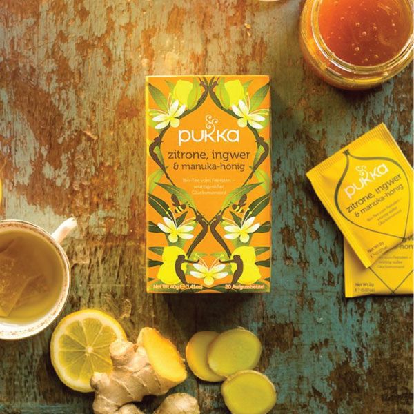 Pukka Tee-Verpackung mit Teebeutel und Zutaten. Zitrone, Ingwer, Honig. Auf Holzoberfläche.