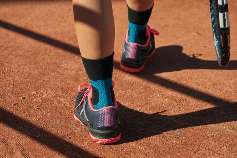 Primo piano dei piedi in piedi su un campo da tennis, con calzini neri Enforma Achilles Support e scarpe sportive. I calzini hanno accenti blu. Una racchetta da tennis è nell'immagine.