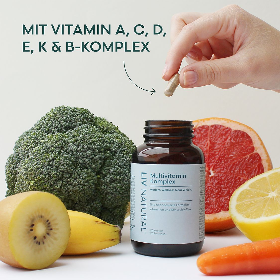 Hand hält Kapsel über Obst und Gemüse. Text: Mit Vitamin A, C, D, E, K & B-Komplex. Flasche mit Kapseln.