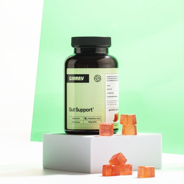 Braune Flasche mit GIMMY Gut Support Gummies. Einige Gummies liegen daneben. Auf weißem Podest, grüner Hintergrund.