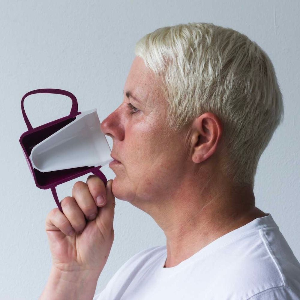 Person trinkt aus einem burgunderfarbenen Becher mit zwei Griffen. Der Becher hat einen weißen Deckel. Die Person hält den Becher mit einer Hand.
