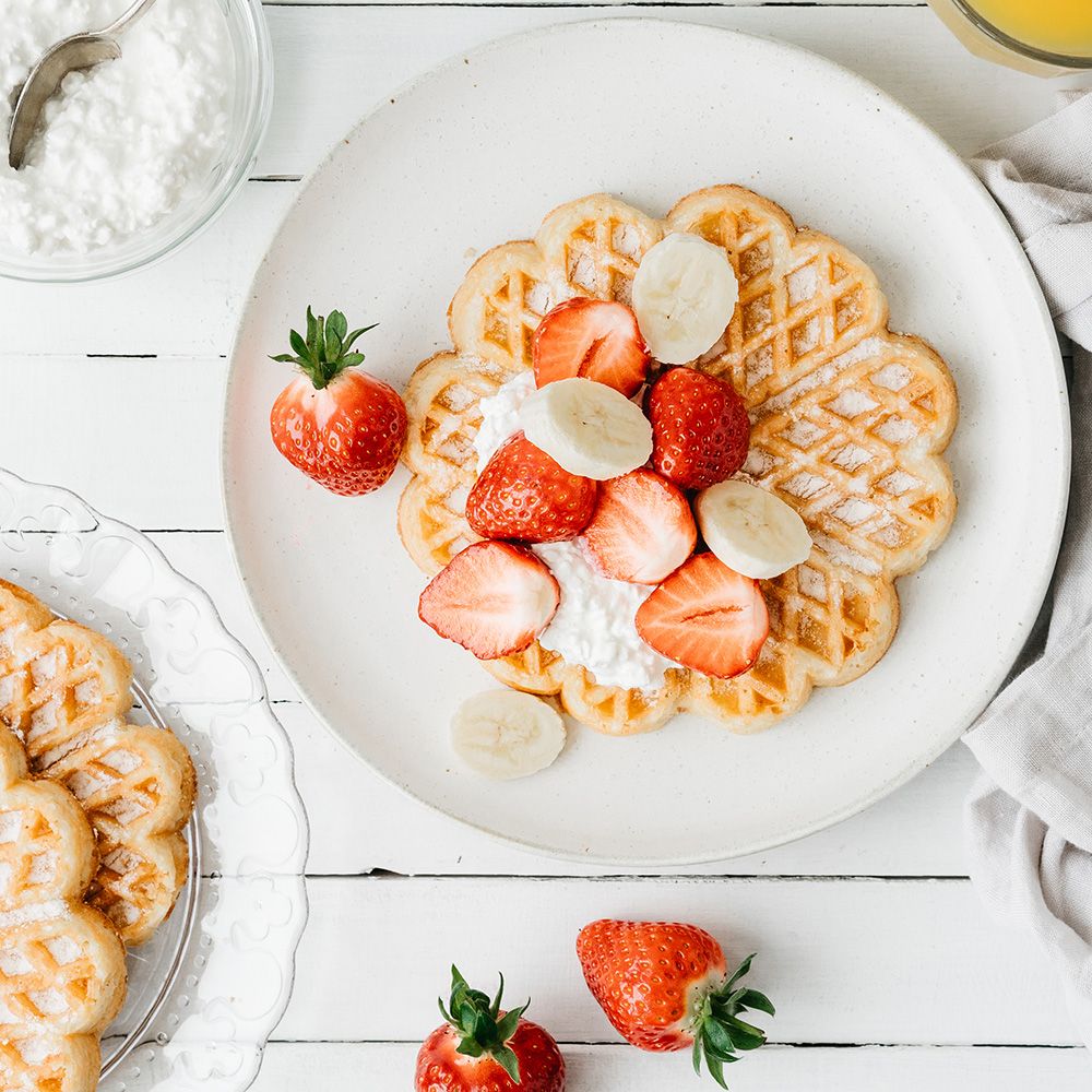 Waffel mit Erdbeeren, Bananen und Creme auf einem Teller. Weitere Waffeln und Erdbeeren liegen daneben. Helle Umgebung.