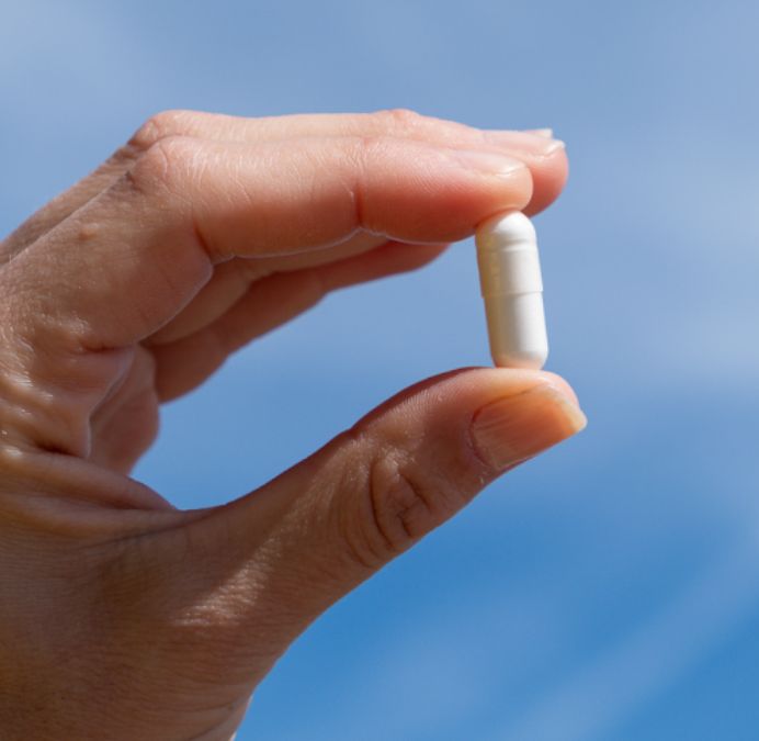 Finger hält eine einzelne weiße Kapsel vor blauem Himmel.