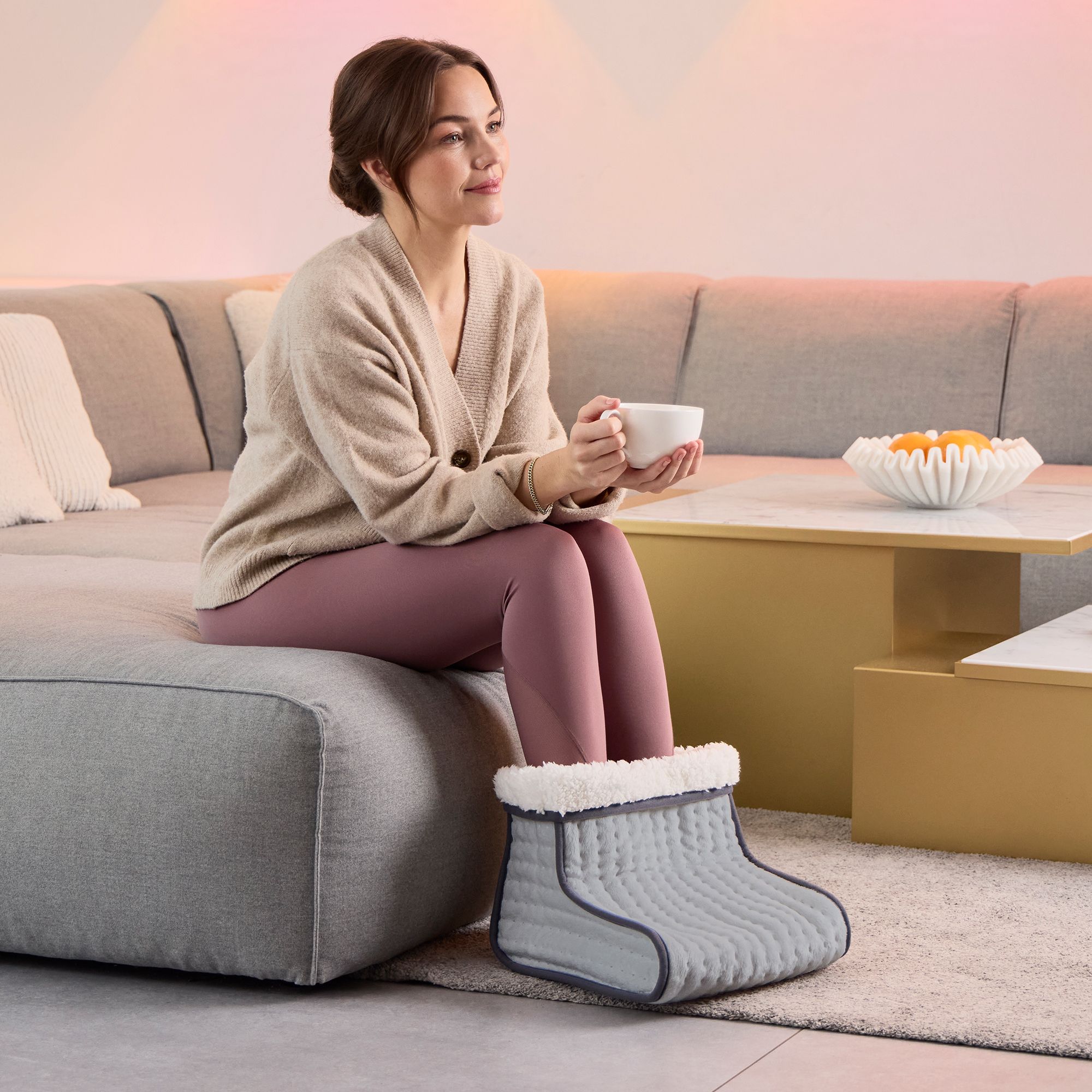 Frau sitzt auf einem Sofa und hält eine Tasse. Ihre Füße stecken in einem grauen Fußwärmer mit weißem Innenfutter.