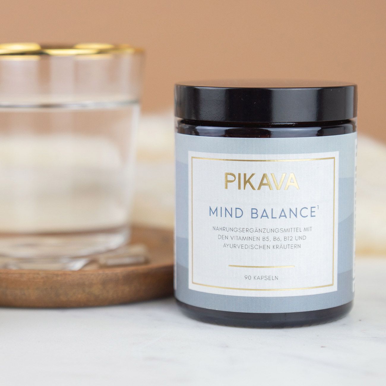 Braune Dose mit schwarzem Deckel. Aufschrift: PIKAVA MIND BALANCE, Nahrungsergänzungsmittel. 90 Kapseln. Glas Wasser.