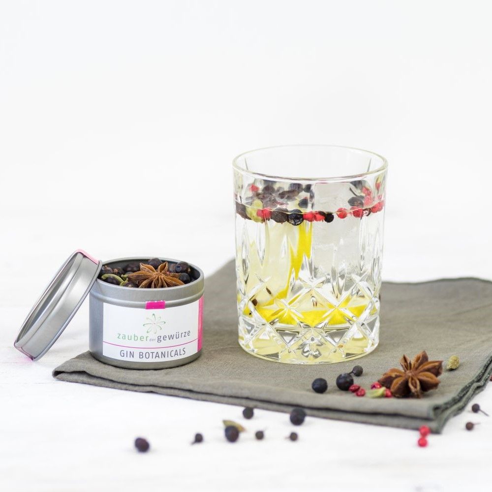 Glas mit Gin und Botanicals. Dose Gin Botanicals daneben. Rosa Etikett mit Schriftzug. Inhalt: Wacholderbeeren, Sternanis, Kardamom.