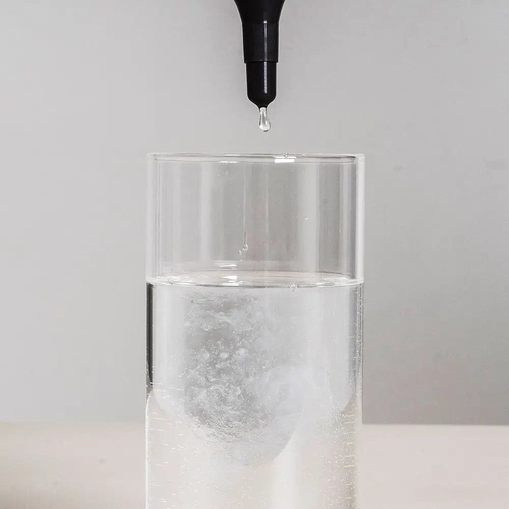 Tropfen aus schwarzem Applikator in ein Glas Wasser. Das Wasser ist klar und leicht trüb.