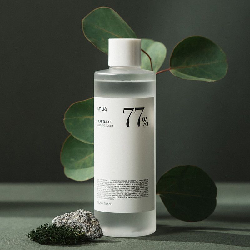 Transparente Flasche mit weißem Deckel und Etikett. Aufschrift: Anua, Heartleaf, 77%. Dekoration: Blätter und Stein.