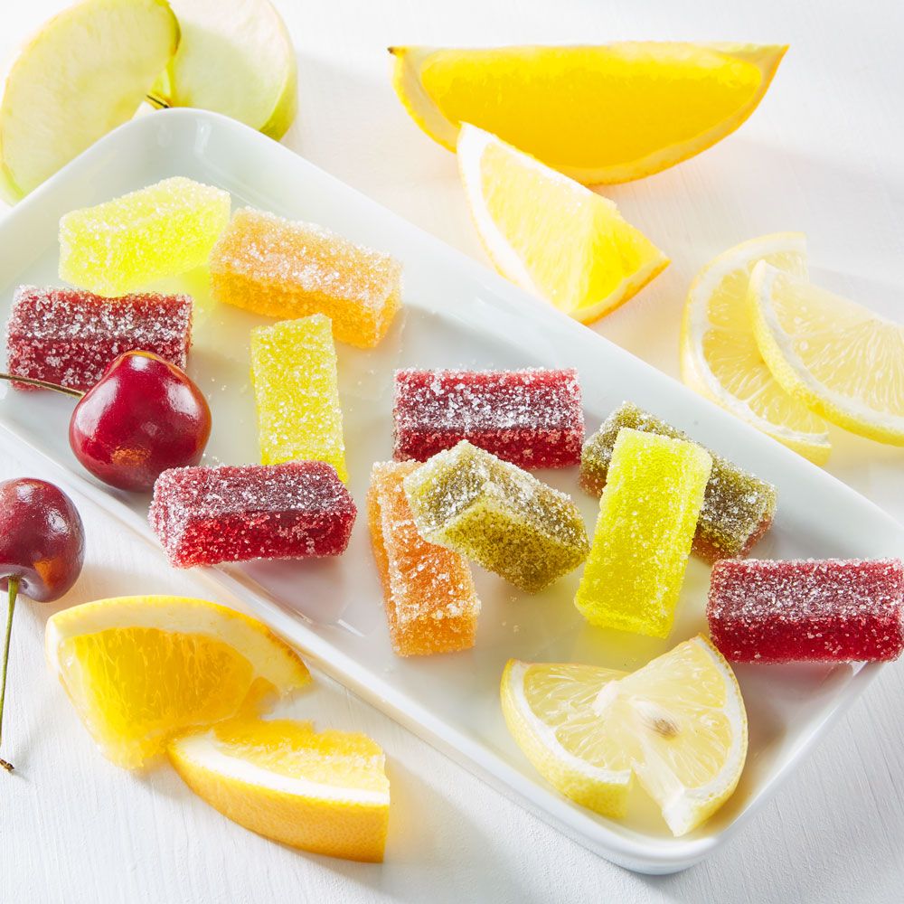 Fruchtmarkgelee-Stücke auf weißem Teller. Verschiedene Farben und Formen. Dekoriert mit Zitronen, Orangen, Äpfeln und Kirschen.