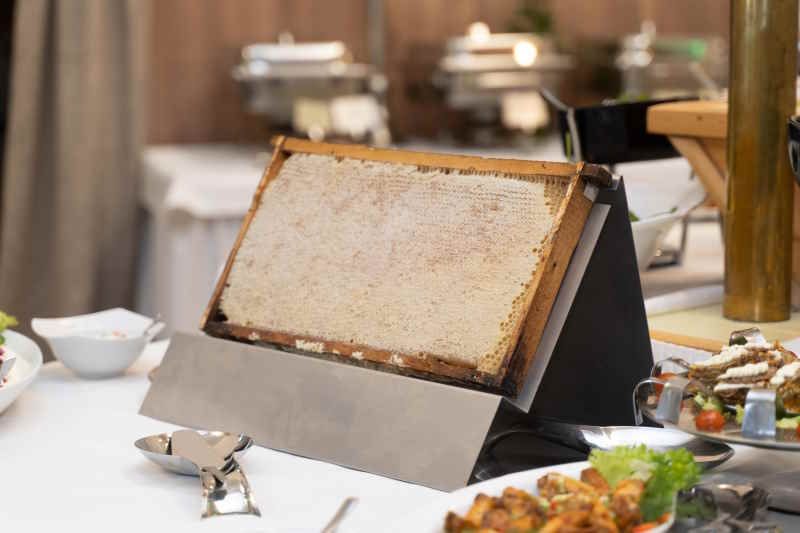 Honigwabe in Holzrahmen auf einem Buffet. Die Wabe ist auf einem Ständer platziert, umgeben von Speisen und Utensilien.