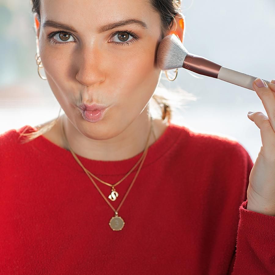 Frau trägt Blush mit einem Pinsel auf. Gesicht zur Kamera gerichtet, Blick nach oben. Roter Pullover.