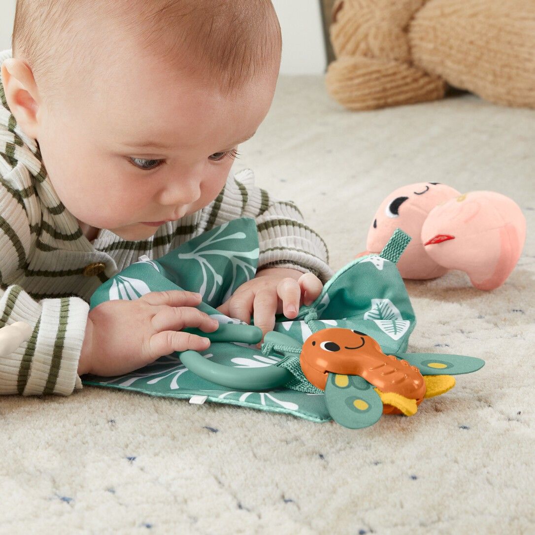 Baby liegt auf einer Decke mit einem Spielzeug. Das Spielzeug hat eine grüne Basis und ein orangefarbenes Element.