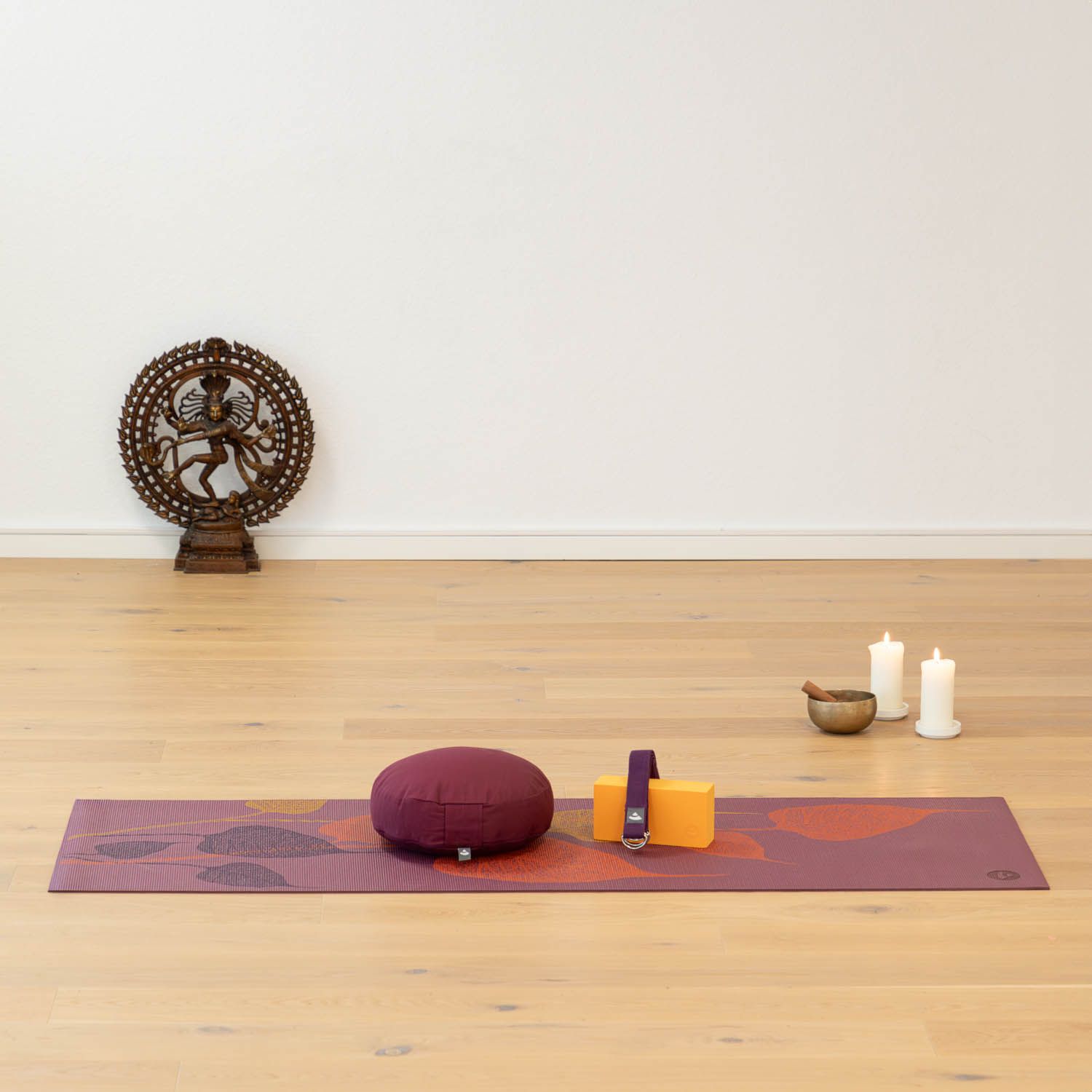 Yogamatte mit Blättermuster, Kissen, Block, Kerzen und Schale. Auf Holzboden. Hintergrund: Wand mit Dekoration.