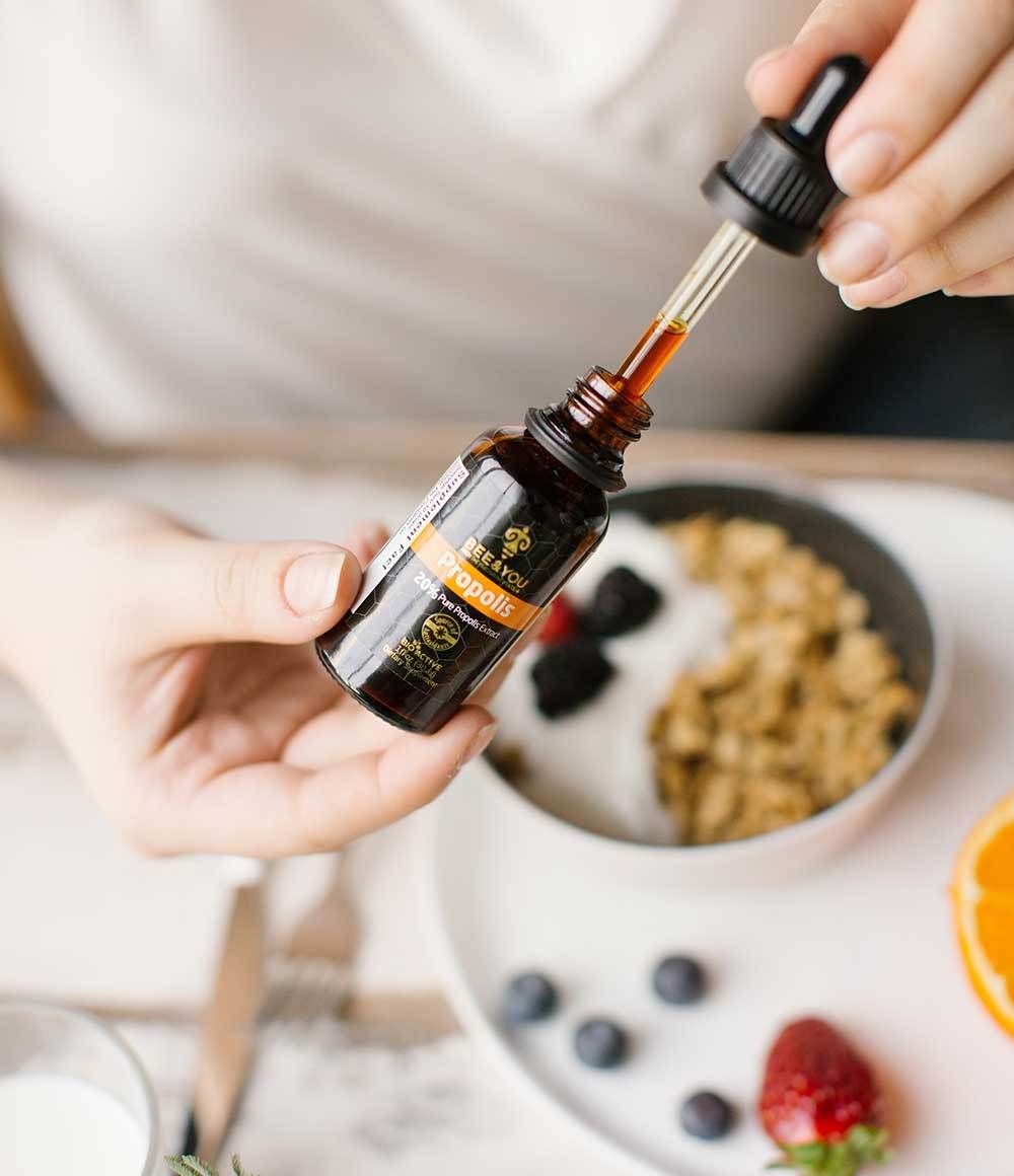 Eine Hand hält eine braune Flasche mit Tropfer über einer Schüssel mit Essen. Auf der Flasche steht "BEE & YOU Propolis".
