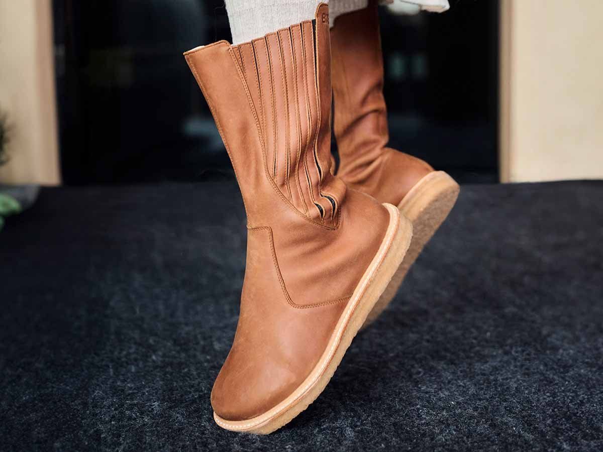Braune Lederstiefel mit breiter Passform. Helle Sohle. Person trägt Stiefel. Schwarzer Teppich.