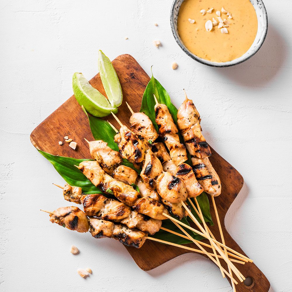 Gegrillte Satay-Spieße auf einem Holzbrett. Limettenscheiben und Soße in einer Schale. Auf weißem Hintergrund.