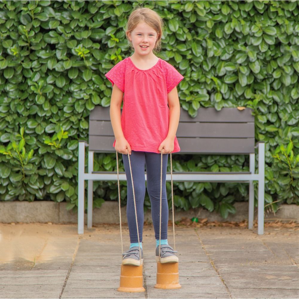 Kind steht auf Laufdosen. Beige Dosen mit Schnüren. Kind trägt rotes Shirt und blaue Hose. Hintergrund: Hecke und Bank.