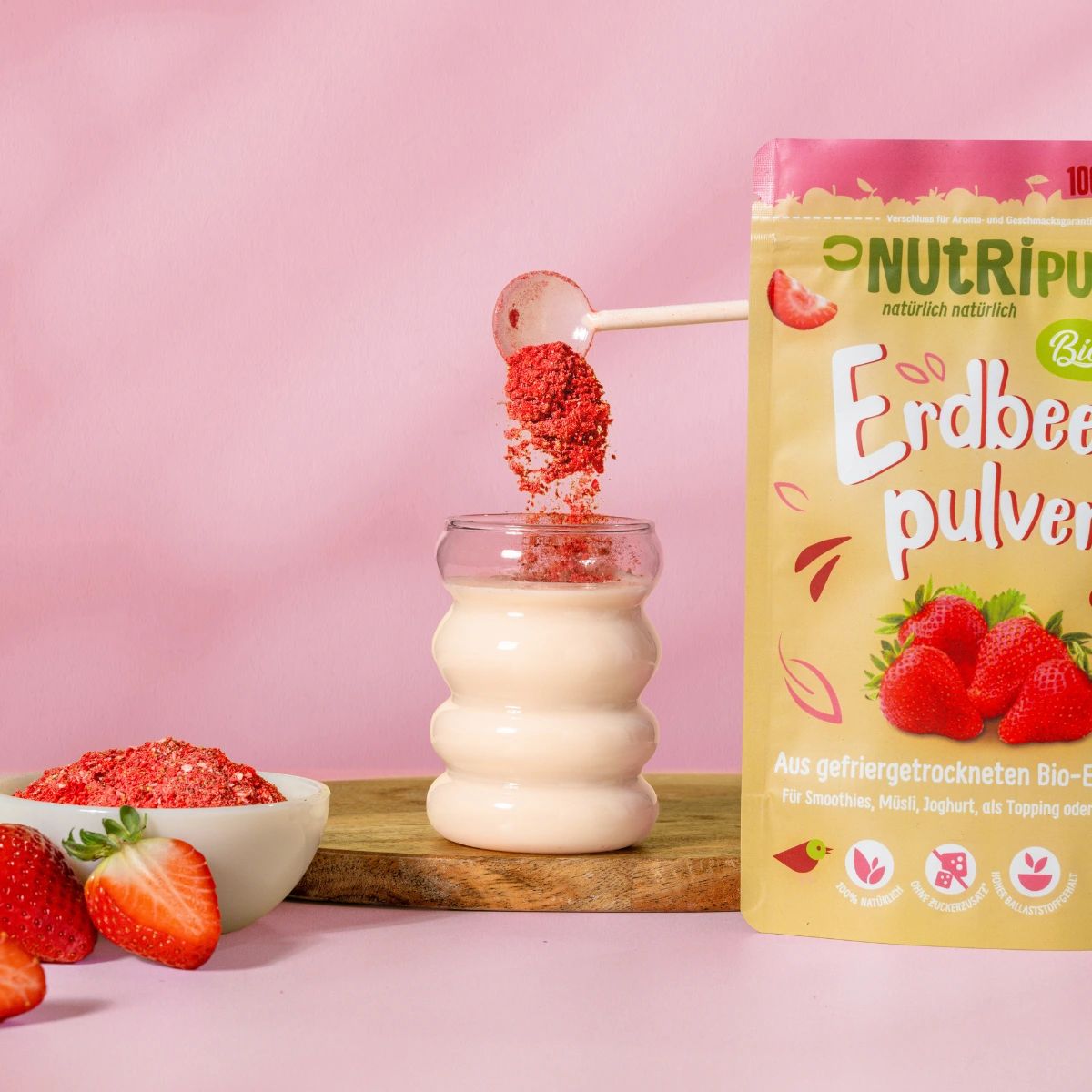 Erdbeerpulver wird in ein Glas gegeben. Daneben Schale mit Pulver, Erdbeeren und Beutel mit Pulver.