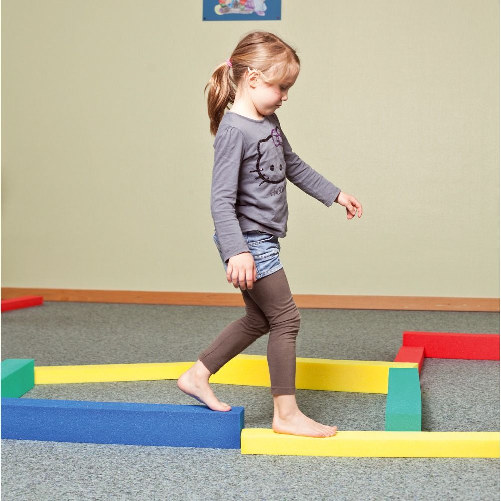 Kind balanciert auf gelben und blauen Schaumstoffbausteinen. Weitere farbige Bausteine liegen auf dem Boden.