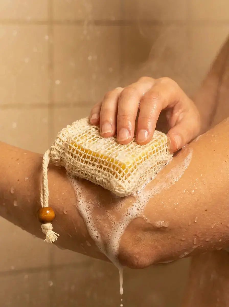 Hand reibt Seife in einem Netzbeutel über den Arm unter der Dusche.