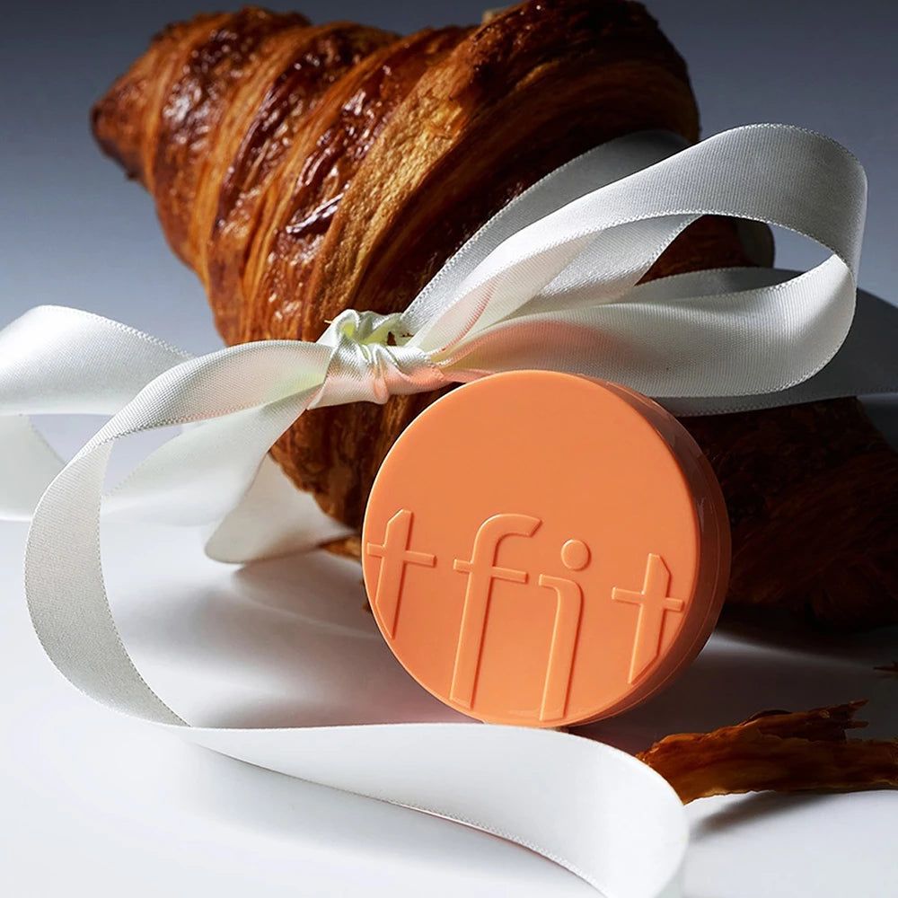 Ein Croissant, gebunden mit einem weißen Band, neben einer runden orangefarbenen Scheibe mit der Prägung 'tfit'.