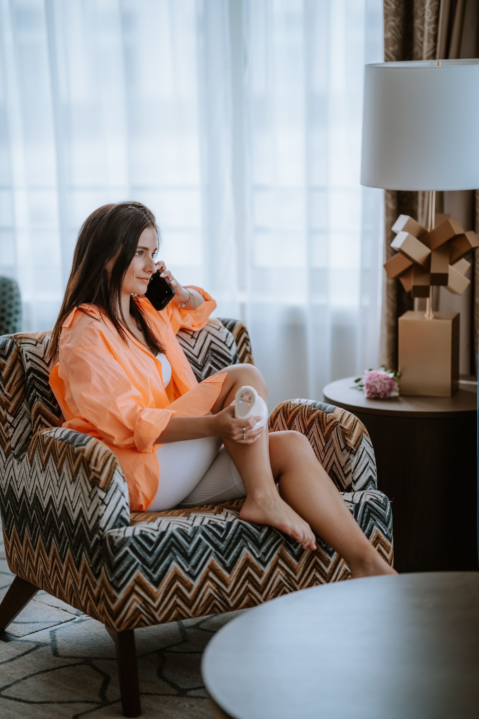 Frau sitzt in einem Sessel und hält ein weißes Massagegerät. Sie telefoniert. Umgebung: Zimmer mit Vorhängen und Lampe.