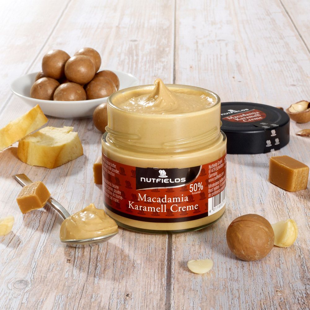 Glas mit Macadamia-Karamell-Creme. Löffel mit Creme. Nüsse, Karamellstücke und weitere Zutaten auf Holzoberfläche.