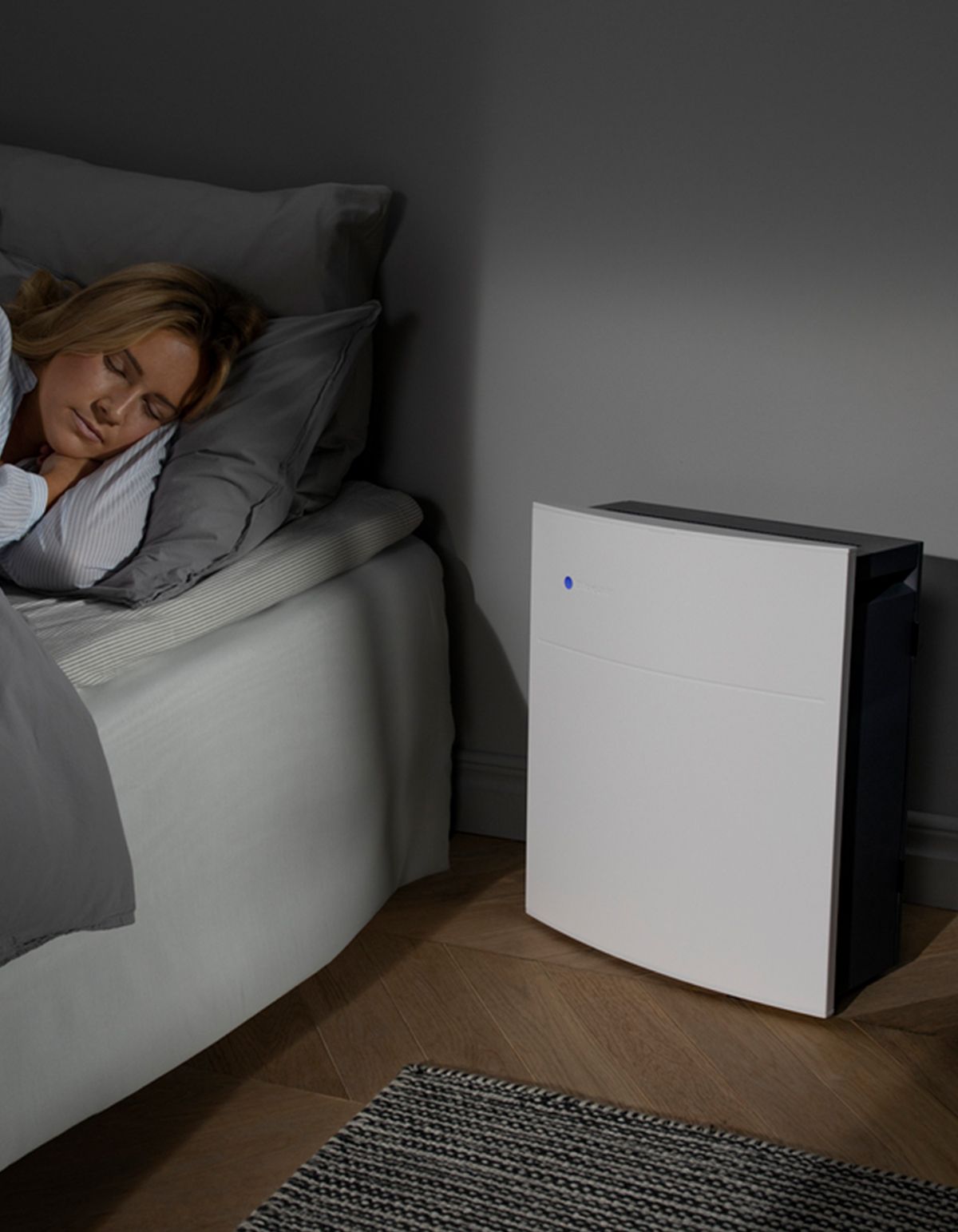 Luftreiniger neben Bett. Frau schläft. Weißes Gerät mit blauer LED. Dunkelgraue Seite.