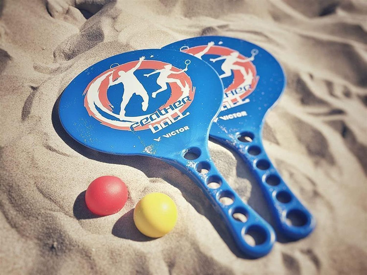 Zwei blaue Federballschläger und zwei Bälle im Sand. Auf den Schlägern steht "Feather Ball" und "Victor".