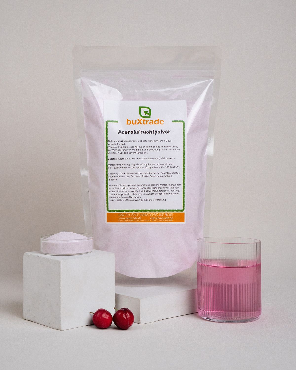 Transparente Tüte, Glas mit rosa Flüssigkeit, Schale mit Pulver, zwei rote Früchte. Aufdruck: buXtrade Acerola Fruchtpulver.