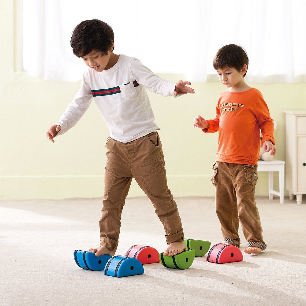 Zwei Kinder benutzen Laufsteine. Blaue, rote und grüne Steine liegen auf dem Boden. Ein Kind balanciert auf blauen Steinen.