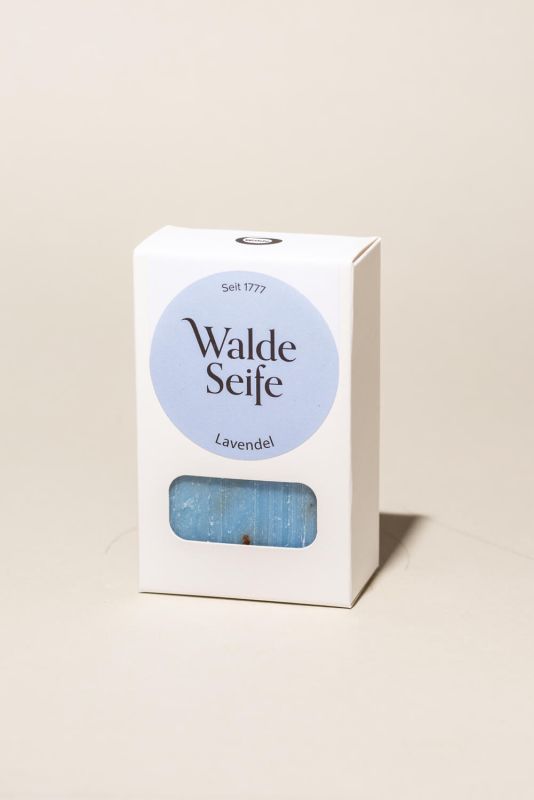 Weiße Verpackung mit blauem Kreis und Text. Sichtbares Seifenstück mit Lavendelblüten. "Walde Seife" und "Lavendel" lesbar.